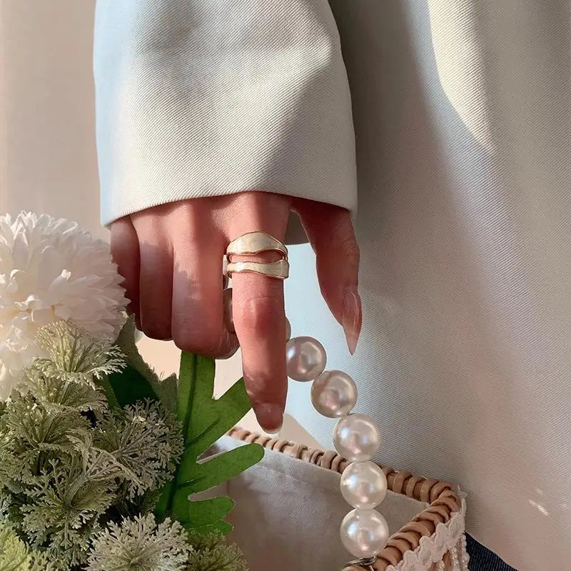 Woman wearing a retro gold open ring with white enamel wavy design, holding a pearl-handle bag and flowers